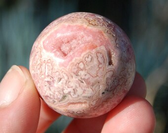 May include: Pink and white sphere of polished gemstone with a natural crystal formation inside. The sphere is smooth and has a swirling pattern.