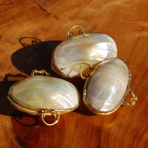 Pode incluir: Três pingentes de formato oval com conchas de madrepérola branca iridescente emolduradas por finas armações de metal dourado. Cada pingente inclui um pequeno aro de metal dourado na lateral para pendurar, com uma superfície lisa e polida.