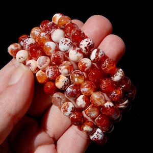 May include: A collection of beaded bracelets featuring round, polished beads in shades of red, orange, and white. The beads have a speckled pattern and are strung together to form multiple bracelets. The bracelets are held in a hand against a black background.