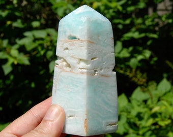May include: A light blue and white layered crystal point, also known as a tower, with a pointed top. The crystal is polished and has a smooth surface.