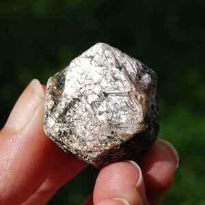 May include: A close-up of a raw, natural mineral specimen. The stone has a multifaceted, crystalline structure with a shimmering, silvery-white surface. Darker, black and brown inclusions are visible throughout the stone, adding contrast. The background is a blurred green.