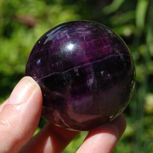 May include: A polished, spherical gemstone with a deep, translucent purple hue. The surface of the sphere displays natural internal fractures, subtle color banding, and reflective highlights that emphasize its smooth, rounded, and solid form.