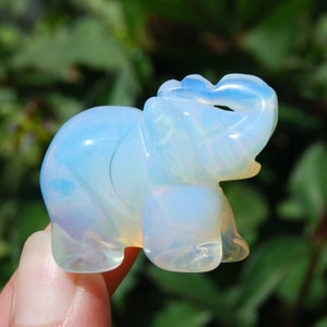May include: A small, white opal elephant figurine. The elephant is carved in a standing position with its trunk raised. The opal has a milky white color with iridescent flashes of blue and green.