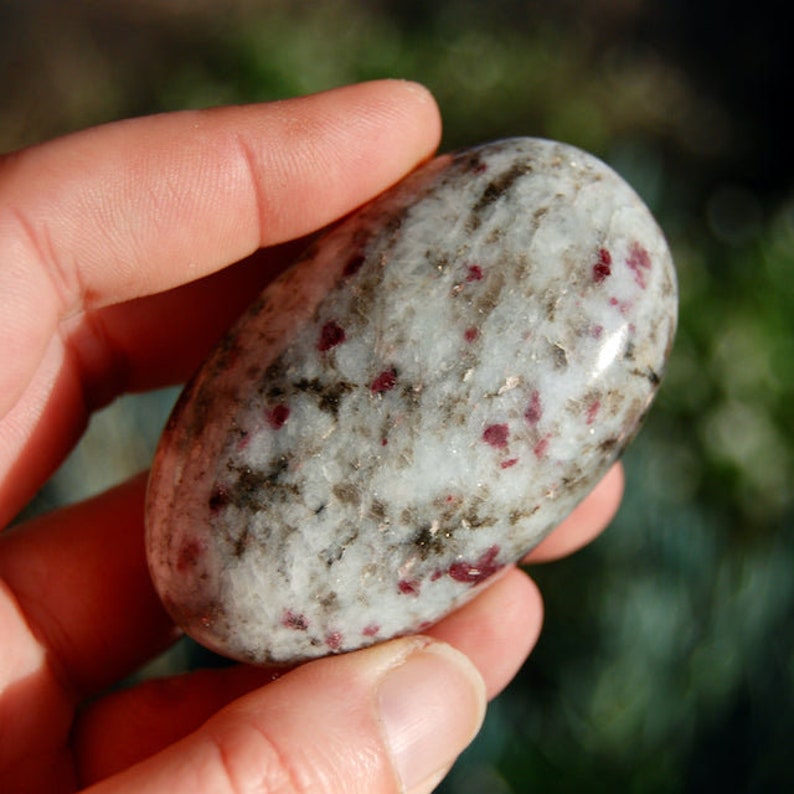 May include: A white, gray, and red tumbled stone with a smooth, polished surface. The stone has a speckled appearance with small red and black inclusions.