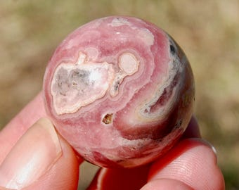 May include: A polished, spherical gemstone with a swirling pattern of pink, white, and gray hues. The stone has a smooth surface and is held in a hand. The gemstone is a decorative item.
