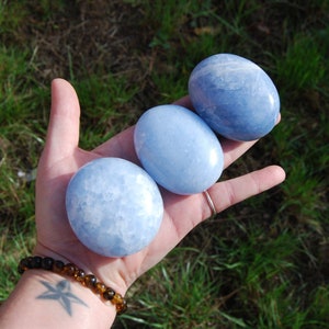 May include: Three smooth, light blue celestite palm stones. The stones are oval shaped and have a white marbled pattern.