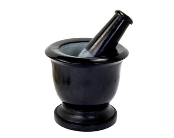 May include: A black mortar and pestle set. The mortar is a small, round bowl with a wide rim and a pedestal base. The pestle is a cylindrical shape with a rounded top. The set is made of a dark, possibly stone material.