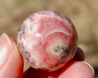 Könnte beinhalten: Eine rosa und weiße Kugel aus poliertem rosa Mangan, auch bekannt als Rhodochrosit, mit einem wirbelnden Muster.