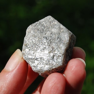 May include: A close-up of a rough, silvery-gray mineral specimen. The rock has a crystalline structure with reflective surfaces, catching the light. The mineral is held in a hand against a blurred green background.
