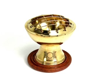 May include: A brass incense burner with a round, grid-covered top and a pedestal base. The base features a triple moon symbol and sits on a round, wooden base. The burner has a polished, golden finish and is designed for burning incense.