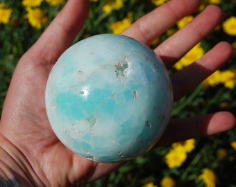 May include: A smooth, light blue and white sphere of Celestite crystal. The sphere has a natural, polished finish and is speckled with small, white inclusions.