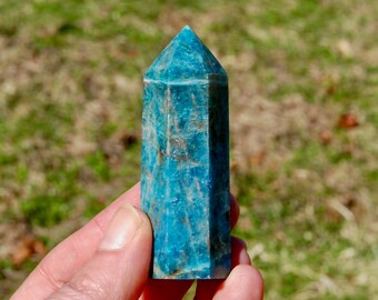 Könnte beinhalten: Ein blauer und grüner Apatit-Kristall, auch bekannt als Turm, wird in einer Hand gehalten. Der Kristall ist facettiert und hat eine spitze Spitze.