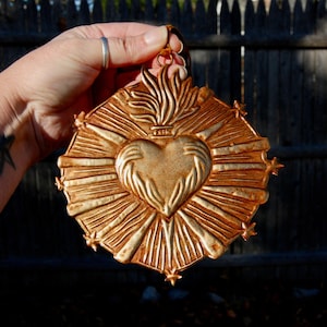Könnte beinhalten: Ein goldfarbenes Metallherz mit Flammen und einer Krone oben, umgeben von einem strahlenden Sonnenstrahlen-Design. Das Herz hat Hände, die sich um es herumschließen.