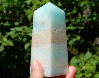 May include: A light blue and white banded aquamarine crystal point, also known as a tower, is held in a hand. The crystal is polished and has a smooth, glassy surface.