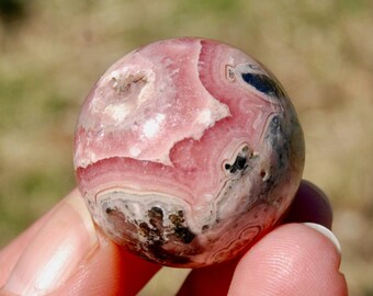 May include: A pink and white banded sphere of polished rhodochrosite, a type of mineral, with black and gray inclusions. The sphere is about 2 inches in diameter.