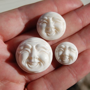 May include: Three white carved bone buttons with smiling faces. The buttons are round and have a raised design. The largest button is about 2 inches in diameter.