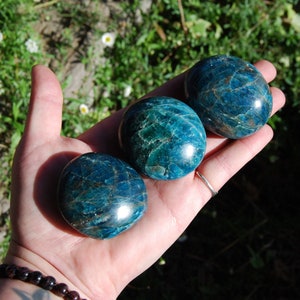 May include: Three smooth, polished blue apatite stones, each with a unique pattern of swirling blue and green hues. The stones are arranged in a row on a hand, showcasing their size and shape.