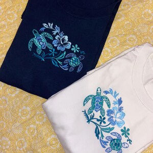 May include: Two t-shirts, one navy blue and one white, are displayed. Each shirt features an embroidered design of sea turtles and flowers in shades of blue and teal. The shirts are folded and rest on a yellow floral patterned surface.