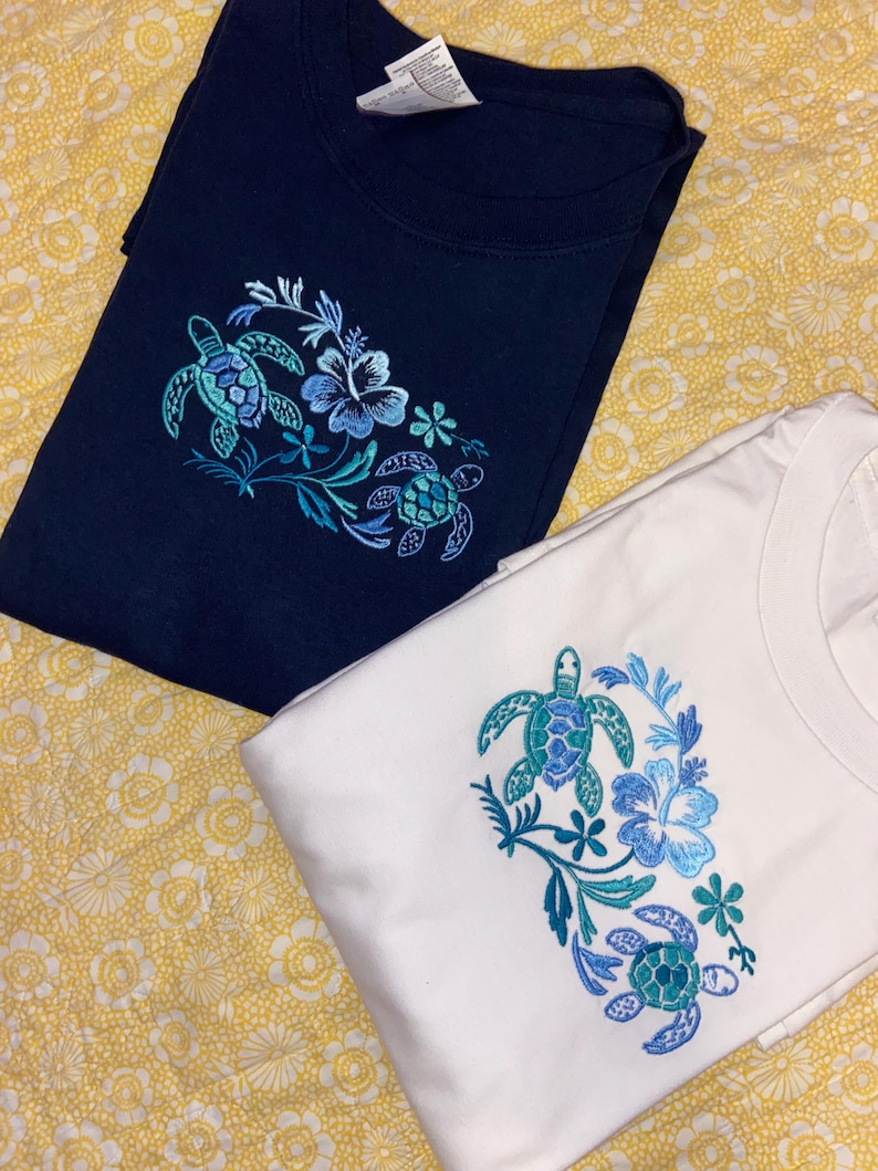 May include: Two t-shirts, one navy blue and one white, are displayed. Each shirt features an embroidered design of sea turtles and flowers in shades of blue and teal. The shirts are folded and rest on a yellow floral patterned surface.