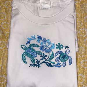 May include: White t-shirt with a crew neck, featuring an embroidered design of two sea turtles and a hibiscus flower in shades of blue and teal. The shirt is folded and displayed on a yellow floral patterned surface.