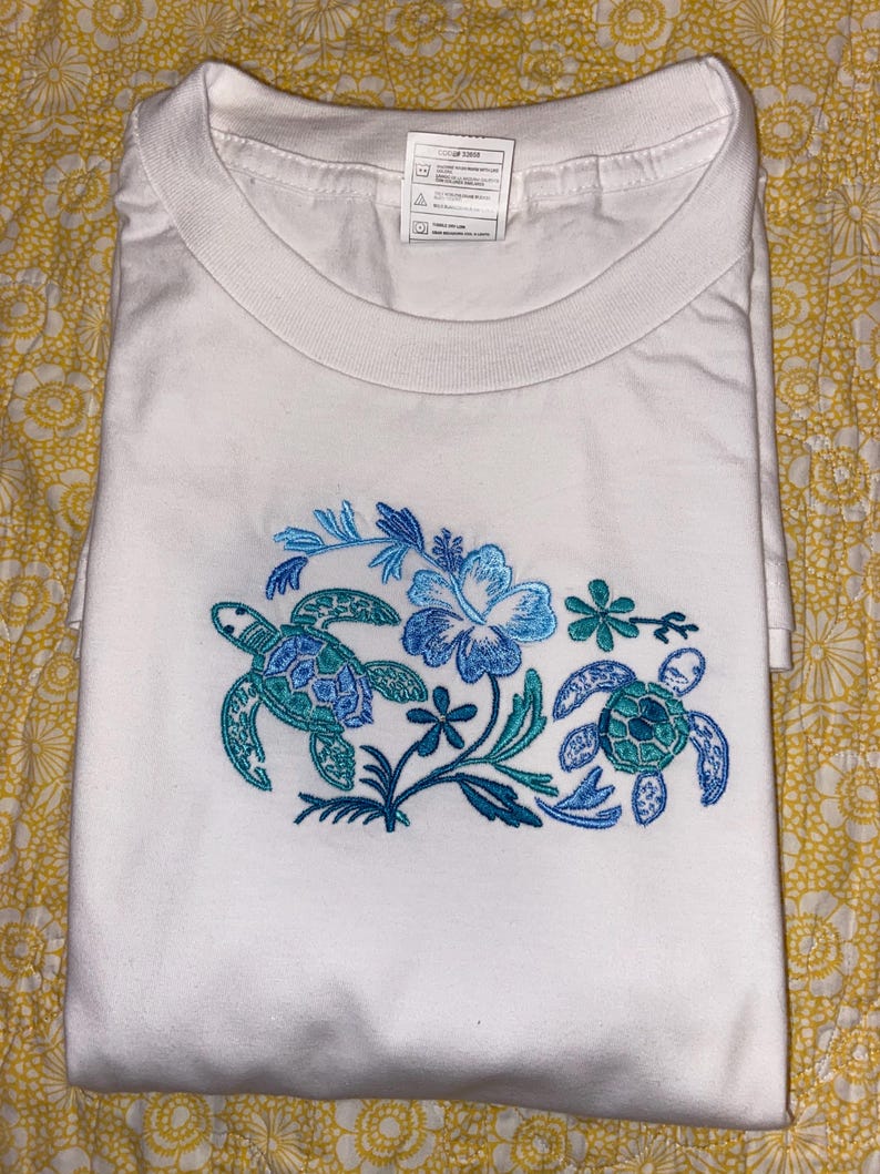May include: White t-shirt with a crew neck, featuring an embroidered design of two sea turtles and a hibiscus flower in shades of blue and teal. The shirt is folded and displayed on a yellow floral patterned surface.
