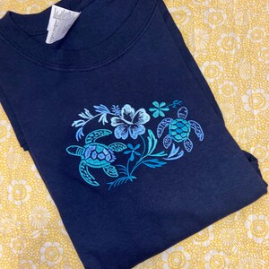 May include: Navy blue t-shirt featuring an embroidered design of two sea turtles and floral elements in shades of blue and teal. The shirt is folded, showcasing the detailed artwork on the front. The background is a yellow floral pattern.
