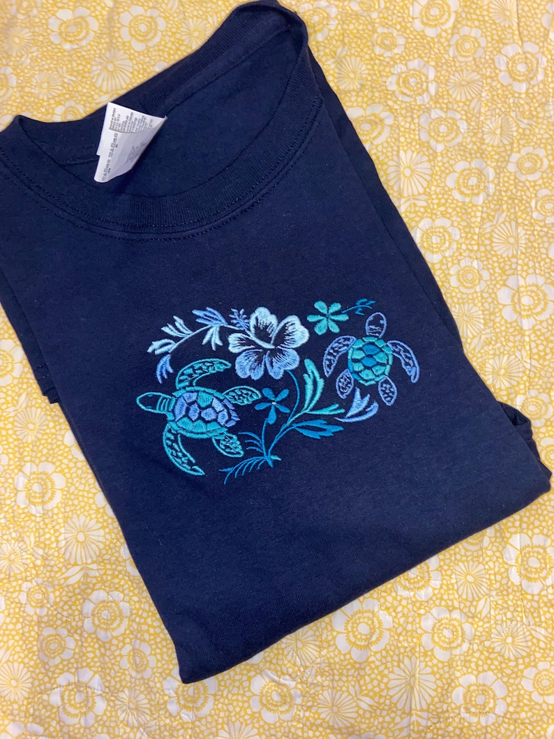 May include: Navy blue t-shirt featuring an embroidered design of two sea turtles and floral elements in shades of blue and teal. The shirt is folded, showcasing the detailed artwork on the front. The background is a yellow floral pattern.