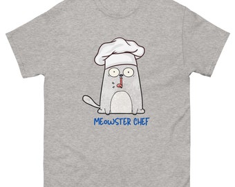 Puede incluir: Camiseta gris claro con un gato de dibujos animados que lleva un gorro de chef blanco. El gato tiene los ojos muy abiertos y saca la lengua. El texto "MEOWSTER CHEF" está impreso en azul debajo.