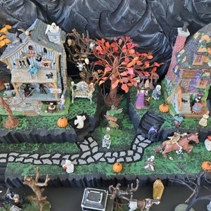 May include: A Halloween village scene with two houses, a witch, a horse and rider, and a graveyard. The houses are decorated with spooky details, including bats, ghosts, and cobwebs. The scene is set on a black base with a green and gray path.