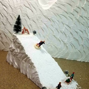 May include: A white styrofoam mountain with a snowy slope. Miniature figures are sledding down the slope. A small evergreen tree is at the top of the mountain.