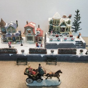 May include: A miniature Christmas village scene with three houses, a horse-drawn carriage, and snow-covered trees. The houses are decorated with lights and have snow-covered roofs. The village is set on a snowy base with a street sign that reads "Main Street".