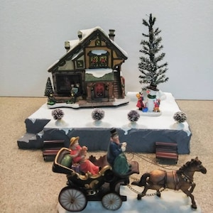 May include: A miniature Christmas village scene with a brown house, a snow-covered landscape, a small evergreen tree, and a horse-drawn carriage with two figures inside.