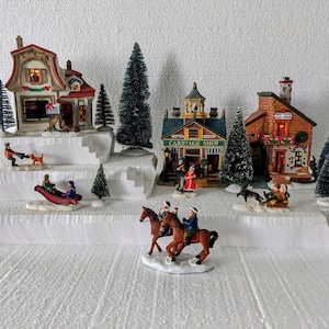May include: A Christmas village scene with miniature houses, trees, and figurines. The houses have red and brown roofs, and the scene includes people sledding and riding horses. The figurines are set on a tiered, snow-covered base.