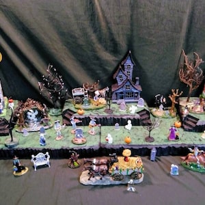 May include: A Halloween village display featuring miniature haunted houses, tombstones, and figurines. The scene includes a full moon, pumpkins, and various spooky characters, creating a festive and eerie atmosphere. The houses are painted in shades of blue, purple, and white.
