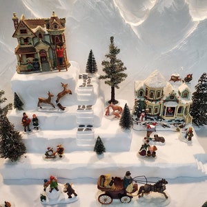 May include: A miniature Christmas village scene with a white snowy base and a multi-level structure. The village features two houses, a horse-drawn carriage, and several miniature figures. The houses are decorated with snow-covered roofs and windows, and the carriage is pulled by a brown horse. The scene is surrounded by miniature trees and other festive decorations.