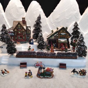 May include: A miniature Christmas village scene with snow-covered mountains, houses, trees, and figures. The houses are decorated with lights and wreaths. The scene includes a sign that reads "Santa's Workshop".