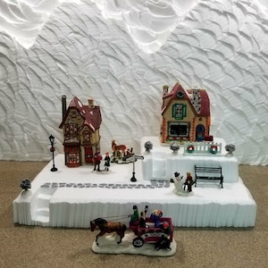 May include: A miniature Christmas village scene with two houses, a horse-drawn carriage, and figures. The houses are decorated with red and white trim and have snow-covered roofs. The carriage is red and gold and is pulled by a brown horse. The figures are dressed in winter clothing and are holding gifts.
