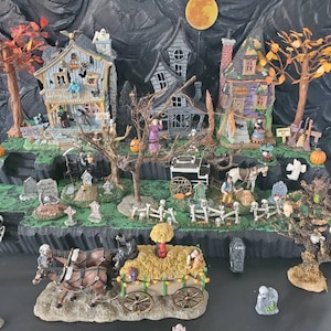 May include: A Halloween village scene with three houses, a graveyard, and a spooky carriage. The houses are decorated with pumpkins, bats, and ghosts. The graveyard has tombstones with spooky messages. The carriage is pulled by two black horses and has a hay bale on the back.