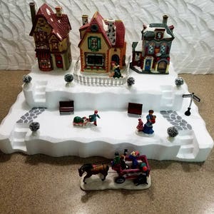May include: A white, snowy village scene with three miniature houses, a horse-drawn carriage, and several figures. The houses are decorated with red and green accents and have white picket fences. The carriage is red and has a white horse.