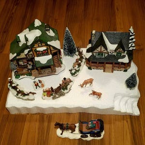 May include: A miniature Christmas village scene featuring two houses, a snowy base, and a horse-drawn sleigh. The houses are decorated with snow-covered roofs and windows. The scene is set on a white base with a snowy landscape.