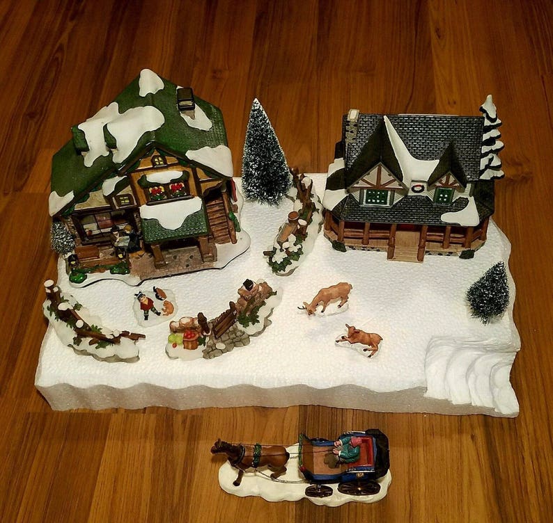 May include: A miniature Christmas village scene featuring two houses, a snowy base, and a horse-drawn sleigh. The houses are decorated with snow-covered roofs and windows. The scene is set on a white base with a snowy landscape.