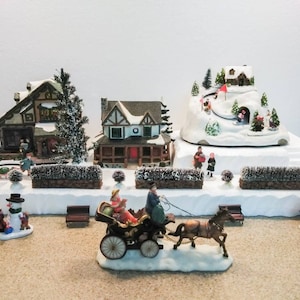 May include: A miniature Christmas village scene with a snowy mountain, a horse-drawn carriage, and three houses. The scene includes a snowman, a street sign, and several miniature people.