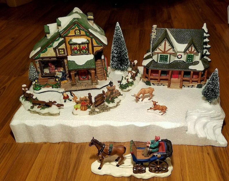 May include: A miniature Christmas village scene featuring two houses, a horse-drawn carriage, and figurines. The houses are decorated with snow, lights, and greenery. The scene is set on a white base with a snowy landscape.