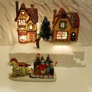 May include: A miniature Christmas village scene with a toy shop, a cottage, and a horse-drawn sleigh with figures. The buildings are lit up with warm white lights. The scene is set on a white base with a snowy effect.
