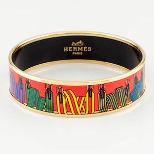 Puede incluir: Un brazalete dorado con un exterior de esmalte rojo con un patrón colorido de equipo ecuestre. El interior es negro y la marca "HERMES PARIS" está impresa en dorado.