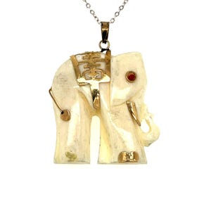 May include: Ivory-colored elephant pendant with gold accents and a red gem, suspended from a silver chain. The elephant has detailed features and a gold emblem on its back. A unique piece of jewelry.