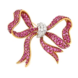 May include: A gold bow brooch, adorned with pink gemstones. The bow's ribbon design is covered in small, round pink stones. The centre of the bow features a cluster of white gemstones. The brooch has a pin closure on the reverse.