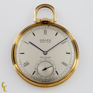 Puede incluir: Un reloj de bolsillo de oro con una esfera blanca y marcadores de hora romanos negros. El reloj tiene una aguja de segundos azul y un subdial más pequeño para los segundos. La esfera del reloj también dice "Gruen Veri-Thin Precision" y "Swiss/Switzerland".