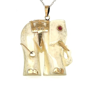 May include: Ivory-colored elephant pendant with gold accents and a red gemstone eye, suspended from a silver chain. The pendant features intricate details, including a gold saddle and decorative elements. A unique piece of jewelry.