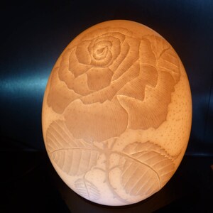 Könnte beinhalten: Eine weiße Straußeneierlampe mit einem geschnitzten Rosenmotiv. Die Lampe ist beleuchtet und strahlt ein warmes Licht aus.